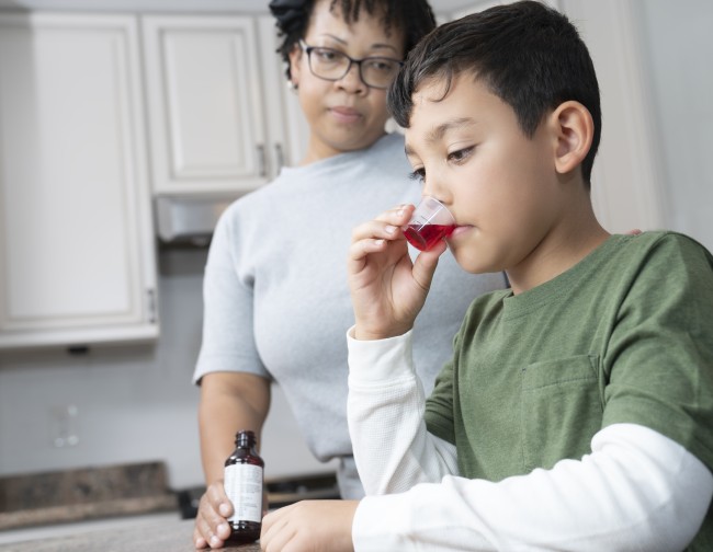 African American/Black woman giving Hispanic/Latinx boy in green shirt liquid medicine