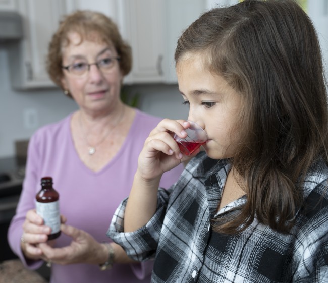 Caucasian/White woman in purple shirt giving ethnically ambiguous young girl liquid medicine