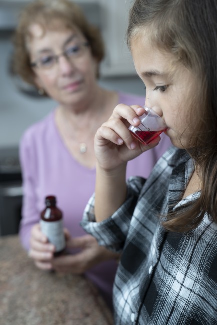 Caucasian/White woman with glasses in purple shirt giving racially ambiguous young girl liquid medicine