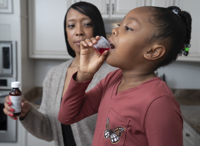 African American/Black woman giving African American/Black girl in red shirt liquid medicine