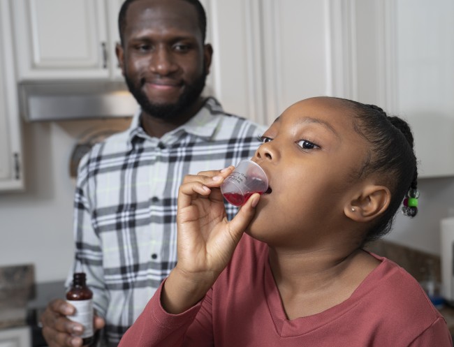 African American/Black man giving African American/Black girl in red shirt liquid medicine