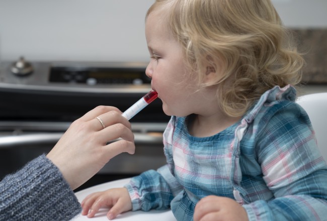 Caucasian/White toddler with blonder hair in blue and pink plaid being given liquid medicine